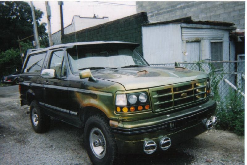 Green camo-painted Ford Bronco with white top and bumper driving lights in a gravel lot