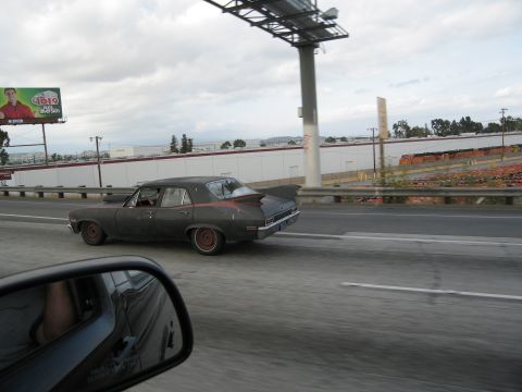 1986 Ford Bronco - A dark sedan with a finned spoiler drives down the highway under an overcast sky.