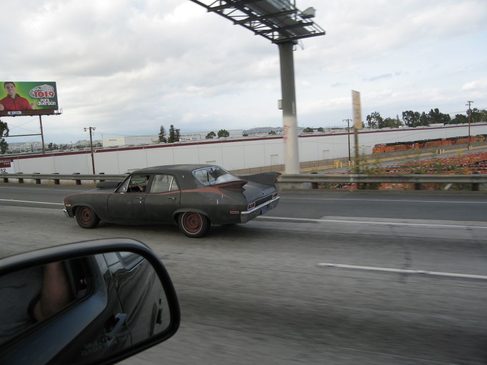 Dark sedan with a red stripe on the rear fender, driving on a highway. The car has a distinctive fin-like spoiler on the trunk