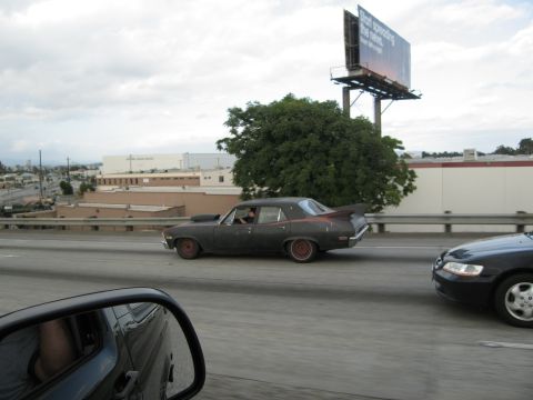 1986 Ford Bronco - A modified dark sedan with a prominent hood scoop and a large rear spoiler drives down the highway.