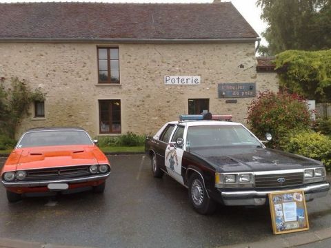 1991 Ford LTD Crown Victoria - Two cars, a police car and an orange muscle car, are parked in front of a building with signs.