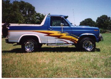 1990 Ford Bronco - This Ford Bronco features a two-tone blue and silver paint scheme with yellow and red flame graphics along the side.