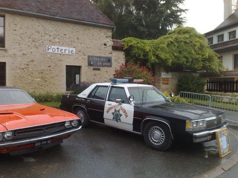 1991 Ford LTD Crown Victoria - A police car and an orange muscle car are parked outside a building with "Potierie" signage.