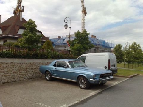 1991 Ford LTD Crown Victoria - A light blue 1967 Ford Mustang coupe is parked next to a white van with construction in the background.