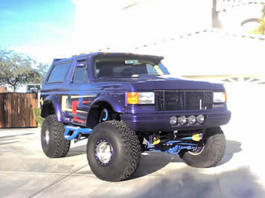 1990 Ford Bronco - Purple Ford Bronco on a tall lift kit parked in a driveway, seen from a low three-quarter front angle with oversized mud tires on beadlock-…