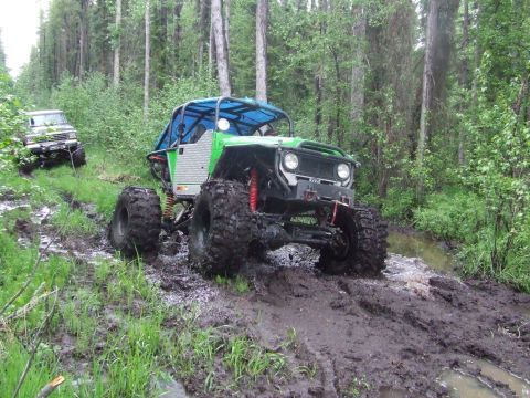 1990 Ford Bronco - Two off-road vehicles traverse a muddy path through a dense forest.