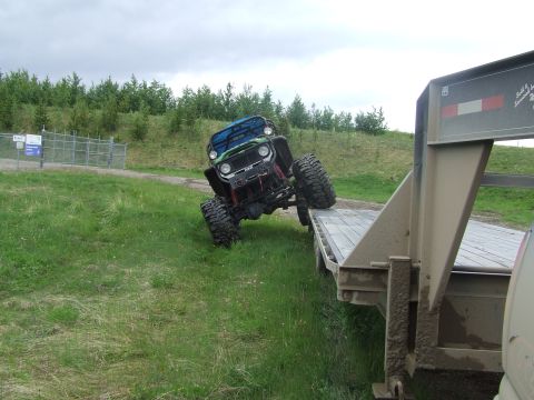 1990 Ford Bronco - A green and blue lifted 4x4 buggy on huge mud-terrain tires flexes its front suspension climbing off a wooden-deck flatbed trailer onto gra…