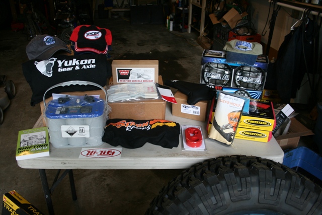 Table with automotive parts including a battery, hats, shirts, and boxes of PIAA lights