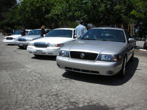 1992 Ford Crown Victoria - Several sedans are parked in a lot under trees, with people standing nearby.