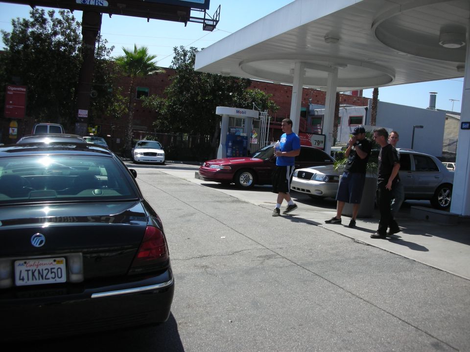 Several cars are at a gas station, including a black sedan in the foreground and a red sedan and silver sedan near the pumps