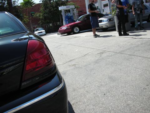 1992 Ford Crown Victoria - Rear view of a black car with a distinctive taillight design, parked at a gas station with other vehicles and people present.
