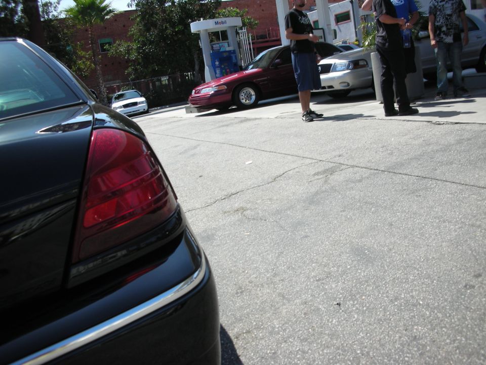 Black car rear quarter panel and taillight. Other cars and people at gas station