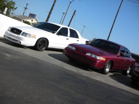 1992 Ford Crown Victoria - Two Ford Crown Victorias, a white one and a maroon one, are parked side-by-side on a sunny day.
