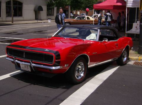 1970 Ford Ranchero - A red convertible muscle car is parked on a street, with other vehicles and people in the background.