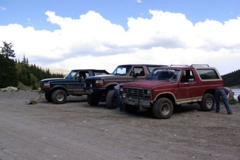 1993 Ford Bronco - Three Ford trucks are parked on a dirt road near a lake, with a person tending to the red truck.