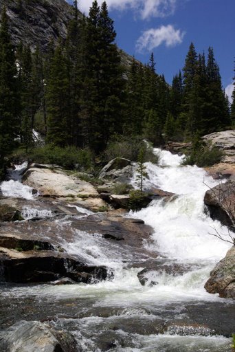 1993 Ford Bronco - A cascading waterfall flows over rocks in a mountainous area with pine trees under a blue sky.