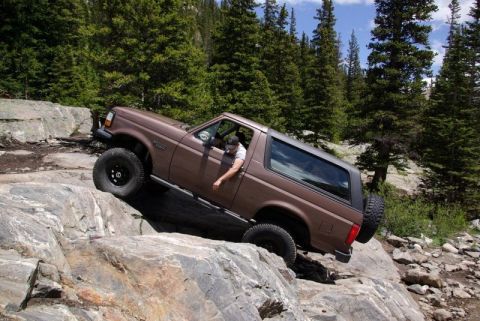 1993 Ford Bronco - A driver navigates a challenging rocky path in a Ford Bronco, surrounded by a dense forest.