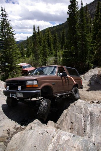 1993 Ford Bronco - A brown Ford Bronco tackles a challenging rocky trail in a mountainous, forested area.