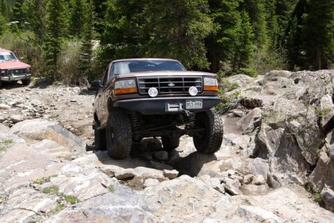 1993 Ford Bronco - A brown Ford Bronco tackles a challenging rocky trail in a wooded mountain area.