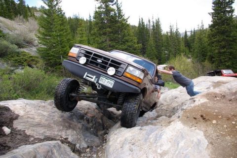 1993 Ford Bronco - A person helps push a modified Ford Bronco over a rocky obstacle on an off-road trail.