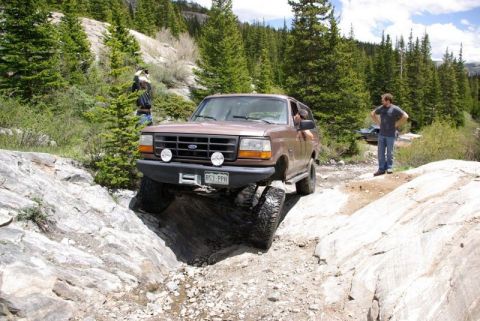 1993 Ford Bronco - A brown 1993 Ford Bronco tackles a challenging rocky incline on a scenic mountain path, with another vehicle visible in the background.