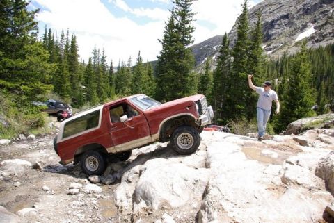 1993 Ford Bronco - A red SUV is shown off-roading on a challenging rocky path in a mountainous, forested area.