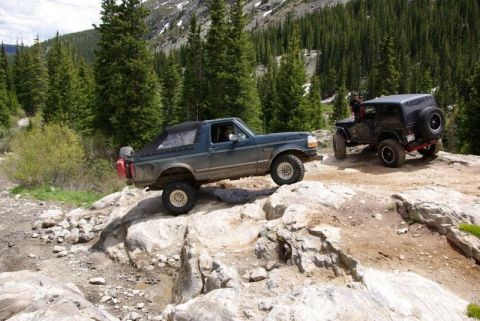 1993 Ford Bronco - Two off-road vehicles tackle a challenging rocky path in a mountainous, forested area.