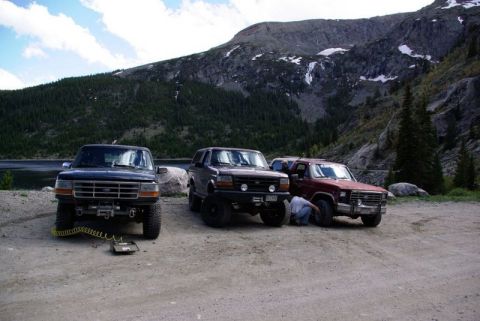 1993 Ford Bronco - Three trucks are parked in a scenic mountain area next to a lake, with one person working on a tire.