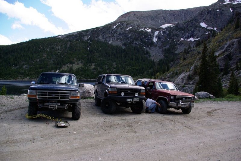 Three trucks parked on a dirt lot with a mountain and lake in the background. One truck is a dark color, one is maroon, and one is black with a winch