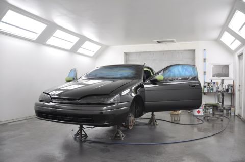 1996 Ford Bronco - A dark-colored sedan is elevated on jack stands inside a brightly lit automotive paint booth.