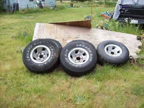 1995 Ford Bronco - Four tires with aluminum rims are displayed on a grassy area next to a wooden structure.