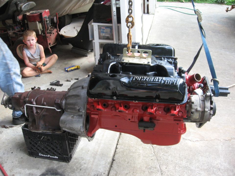 Engine and transmission assembly suspended by a chain, with a child sitting on the floor nearby