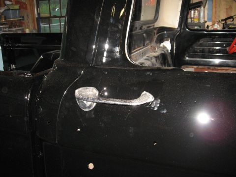 1966 Ford F-100 - Close-up of the chrome pull-style door handle on the freshly painted black door of a pickup cab inside a workshop, with sanding dust and sp…
