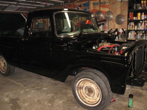 1966 Ford F-100 - A black pickup truck is parked in a garage, with its hood open revealing the engine components.