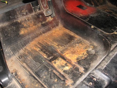 1966 Ford F-100 - Looking down into the stripped cab floor of a pickup with the seat and carpet removed, showing a ribbed rubber floor mat over rusty, patche…