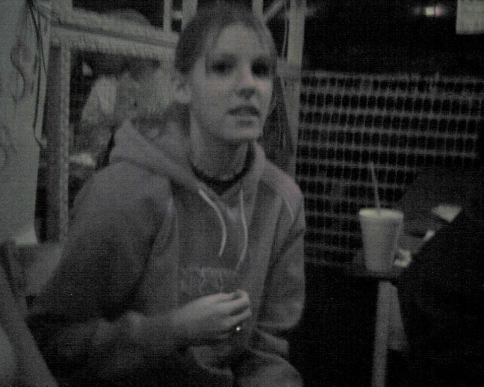 Young woman in a hoodie holding a drink, with a checkered fence and a cup in the background