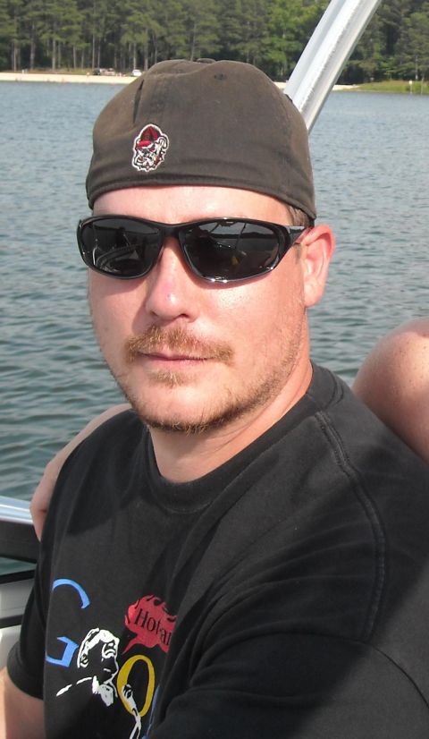1979 Ford F-150 - A man wearing sunglasses and a baseball cap is on a boat with a body of water and trees in the background.