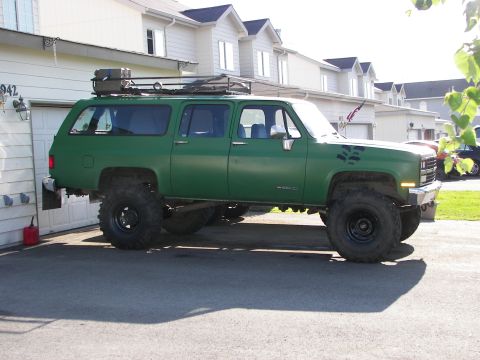 1990 Chevrolet Suburban - This lifted green SUV features a roof rack and large off-road tires, parked in front of a residential garage.