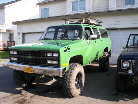 1990 Chevrolet Suburban - This lifted green SUV has large off-road tires and a roof rack, parked in front of a residential garage.