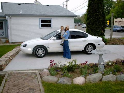 1997 Ford Taurus - A woman and child stand beside a white sedan in front of a house.