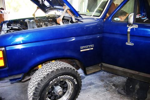 1989 Ford Bronco - A blue Ford Bronco is shown with its hood up, revealing the engine bay. The driver's side door is open.
