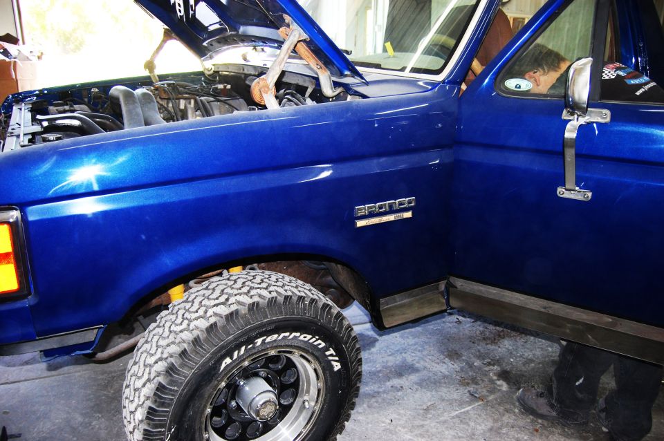 Blue Ford Bronco with hood open, showing engine. Door is open, person inside