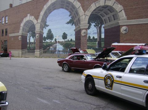 1970 Ford Ranchero - A police car and several classic cars are parked in front of a building with a large mural.