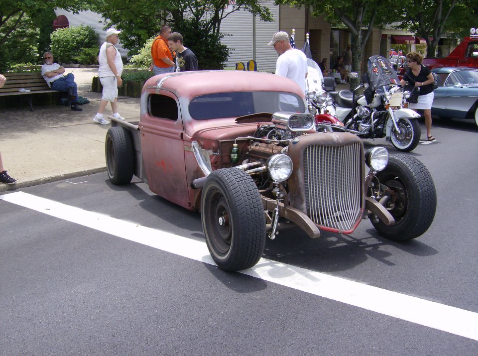 Rat rod pickup with exposed engine and large grille, parked on street with motorcycles and other cars nearby