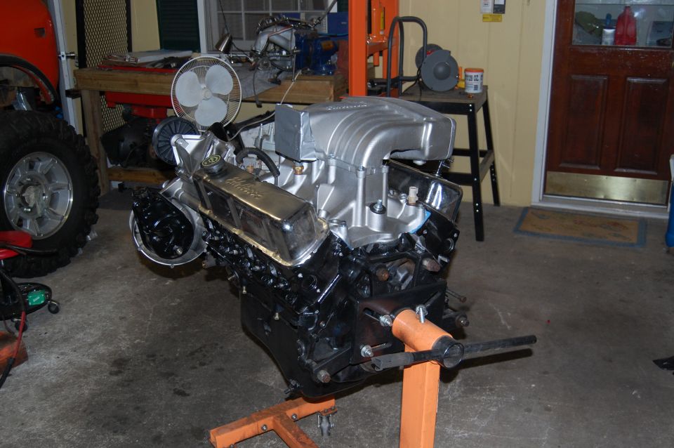 Engine on an orange stand, with a large silver intake manifold and chrome valve covers. A tire and part of an orange vehicle are visible to the left