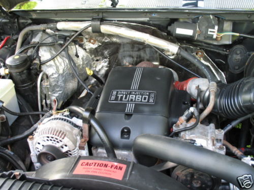 Engine bay with black "DIRECT INJECTION TURBO DIESEL" cover, alternator, and red "CAUTION-FAN" sticker