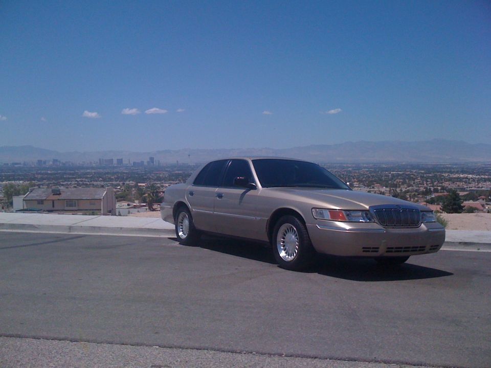 Gold Mercury Grand Marquis sedan parked on a hill overlooking a desert city skyline
