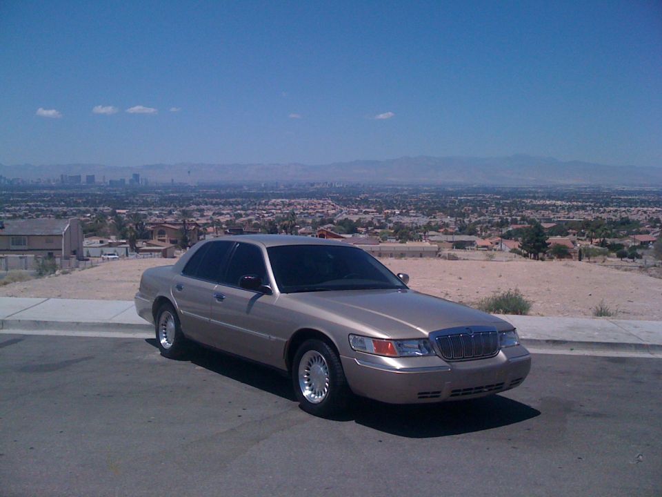 Gold Mercury Grand Marquis sedan parked on a street with a city skyline in the background