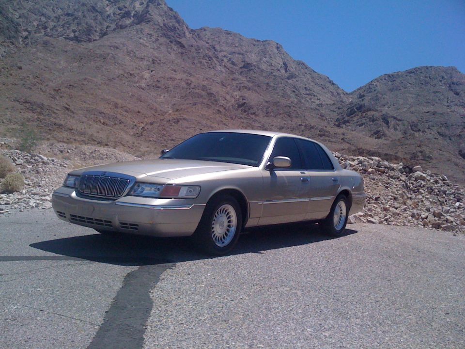 Gold Mercury Grand Marquis parked on a rocky roadside with mountains in the background