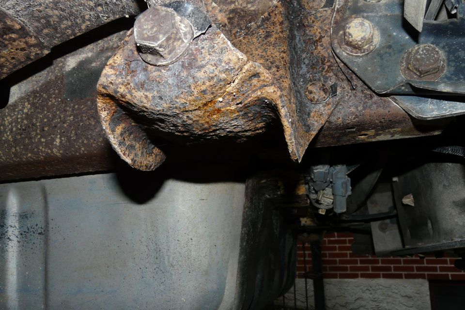 Heavily rusted metal frame and suspension components of an undercarriage. Visible bolts and brackets show significant corrosion